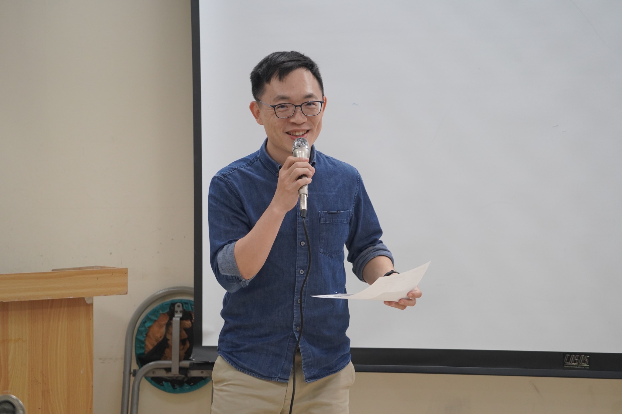 Ji-Jia Ding smiling while holding a microphone and speech notes at a club meeting.